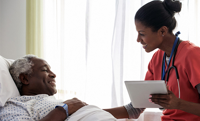 A Black nurse talks to a senior patient in a hospital bed.