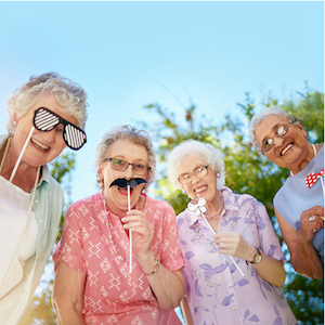 Four senior women smile outside while playing with costume mustaches and glasses