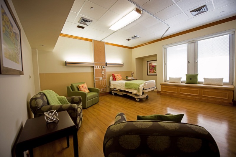 A patient room with a bed, several armchairs and two sunny windows