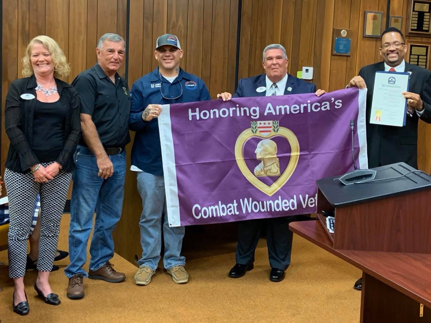 The group stands holding the Purple Heart flag, and the mayor holds a proclamation