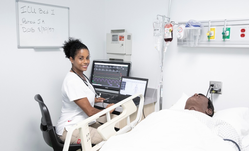 A nursing student sits at a computer near the manikin in a hospital bed