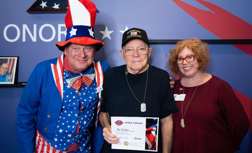 A veteran with a certificate of appreciation in front of the Veterans Wall, flanked by two staff members