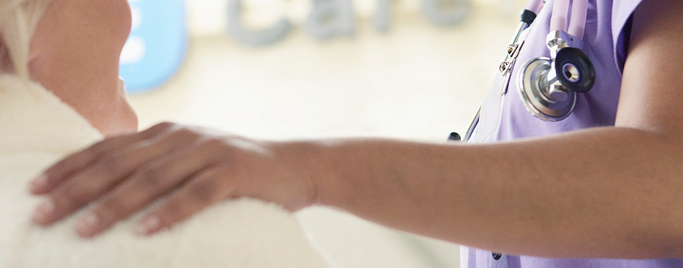 A woman wearing scrubs and a stethoscope lays a hand on a patient's shoulder.