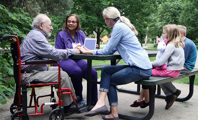 Un paciente en una silla de ruedas se sienta en una mesa de picnic con sus parientes y un miembro del equipo de VITAS