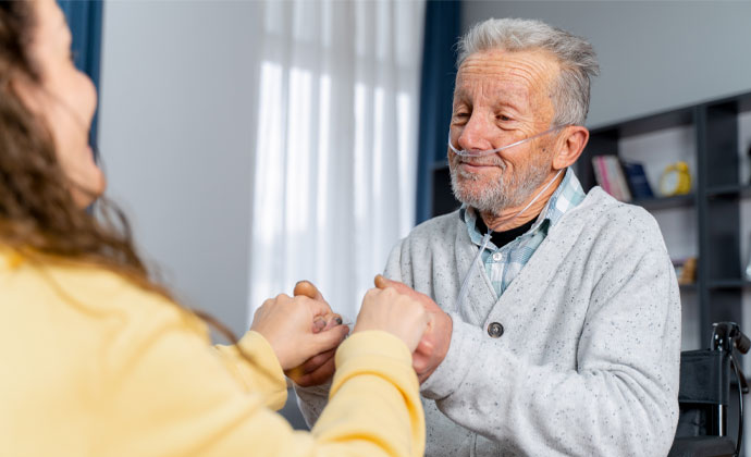 An older man sitting in a chair and wearing a nasal cannula smiles and holds the hands of his daughter
