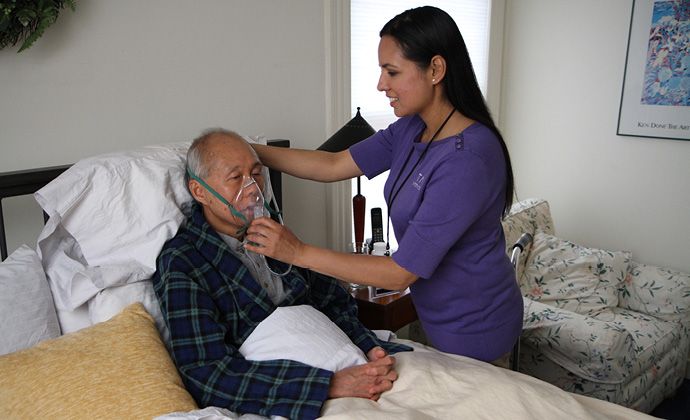 Un miembro del equipo de VITAS ayuda a un paciente con su máscara de oxígeno sentado en una cama