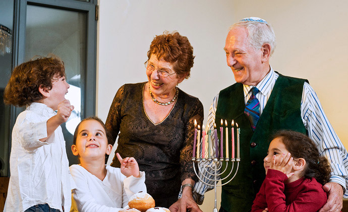 Nakangiti ang isang lolo at lola sa kanilang tatlong apo sa paligid ng isang may sinding menorah