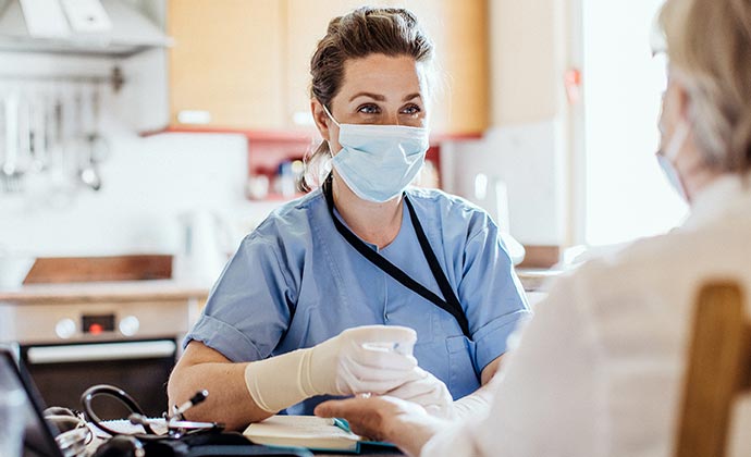 A nursing assistant wearing a mask sits and checks up on her patient