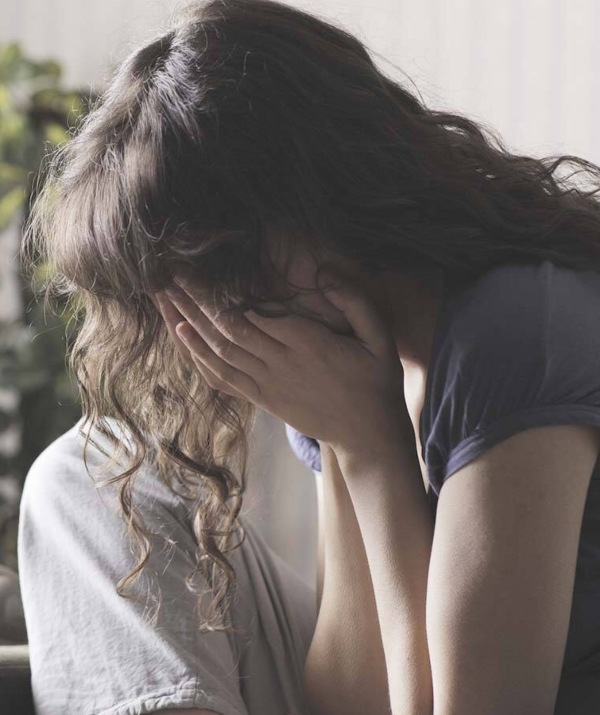 A teenage girl, with her head down, hugs her knees as she sits on a sofa