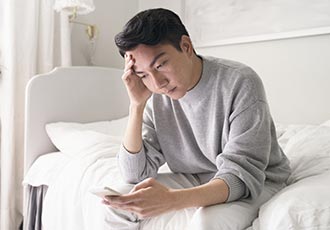 Một người đàn ông đau buồn ngồi ở mép giường và nhìn vào chiếc điện thoại thông minh