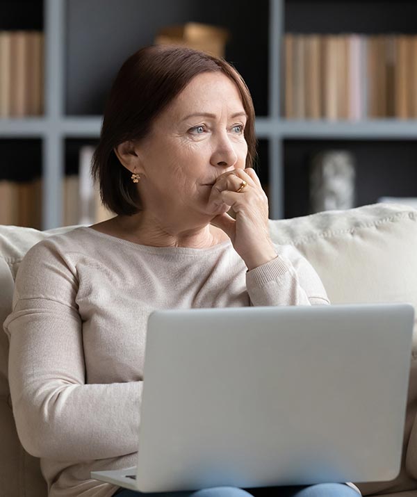 Isang babae na malayo ang tingin habang nakaupo sa sofa at may laptop sa kanyang kandungan