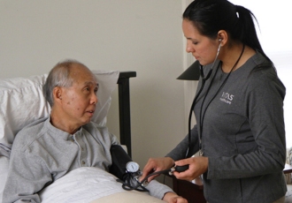A VITAS nurse uses a stethoscope to check on a patient as he sits up in bed at home