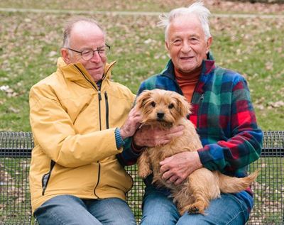 An older gay couple sits on a park bench with their small dog