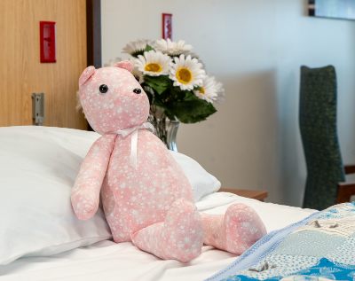 A pink teddy bear near the pillow on a bed, with a vase of white daisies in the background