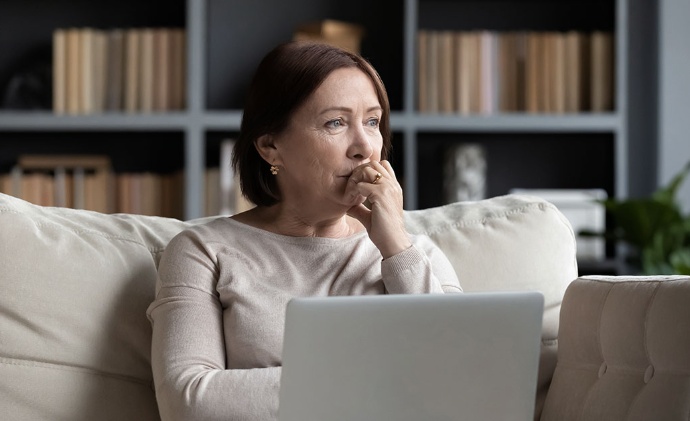 A woman sits on a sofa with a laptop, looking into the distance