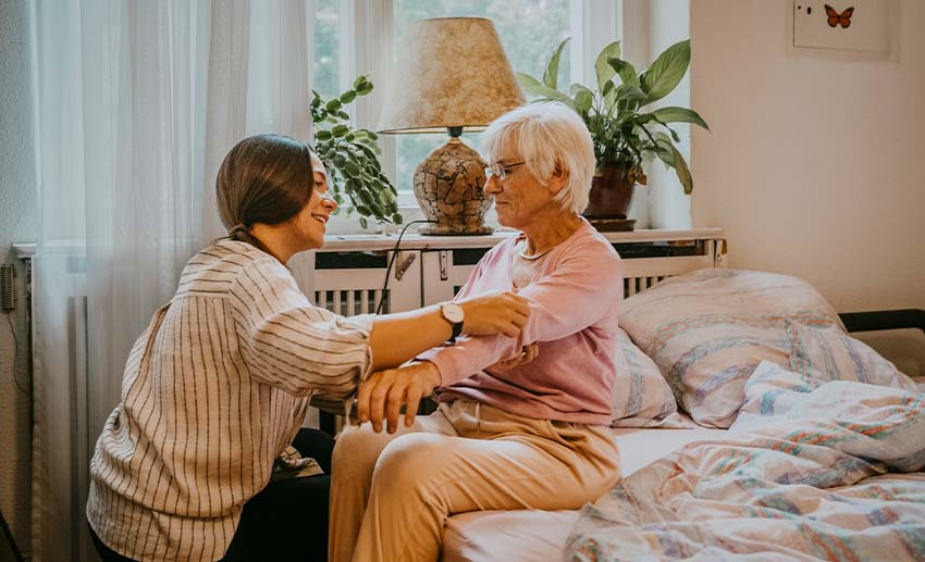 Một người phụ nữ quỳ gối kiểm tra huyết áp cho một người cao tuổi đang ngồi trên giường