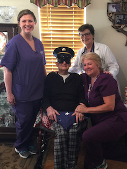 100 year old veteran sits surrounded by staff with his honorary flag