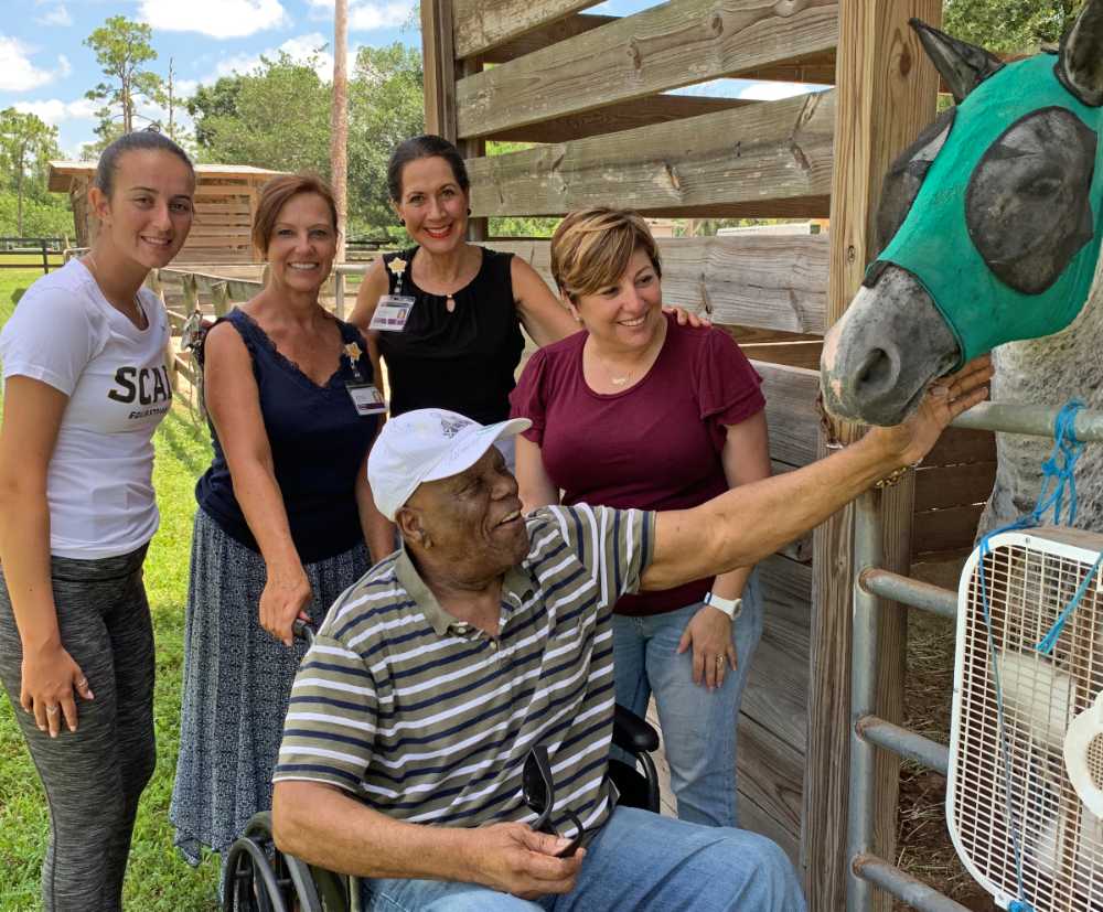 The team stands behind Roy as he touches Colin, a grey male horse, at a stable