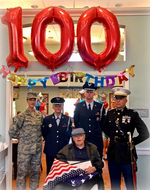 Wynn and the Honor Guard pose under an archway decorated with a banner spelling out Happy Birthday and three large balloons for 1-0-0