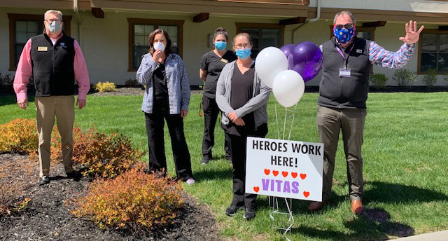 The Kansas City team, wearing masks, cheer on workers at a healthcare facility with balloons, smiles and waves