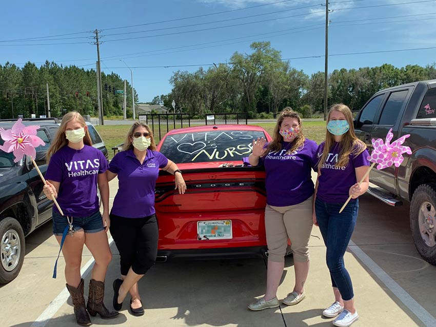 Four VITAS team members, wearing masks, hold giant pinwheels as they pose behind a Dodge Charger with "VITAS hearts Nurses" on its rear window