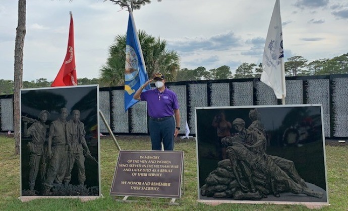 Fred salutes in front of three flags, a memorial plaque and two pieces of artwork commemorating those who served in Vietnam