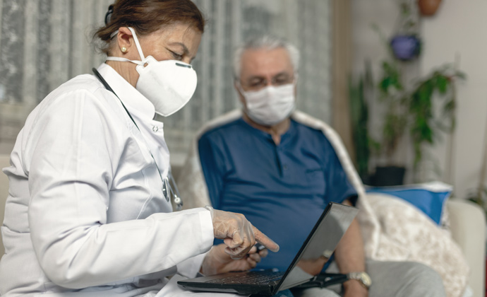 A healthcare professional wearing a protective mask and gloves explains medical information to a hospice patient at home