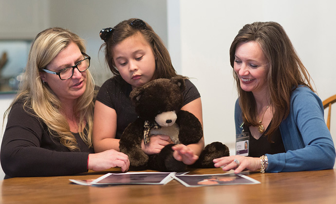 A VITAS chaplain talks with a young girl and her mother at their kitchen table, while the girl holds a teddy bear