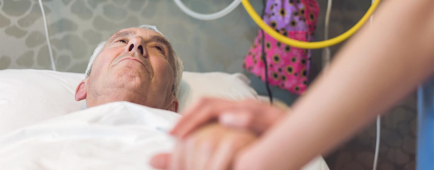 A patient lies in a hospital bed holding the hand of his caregiver