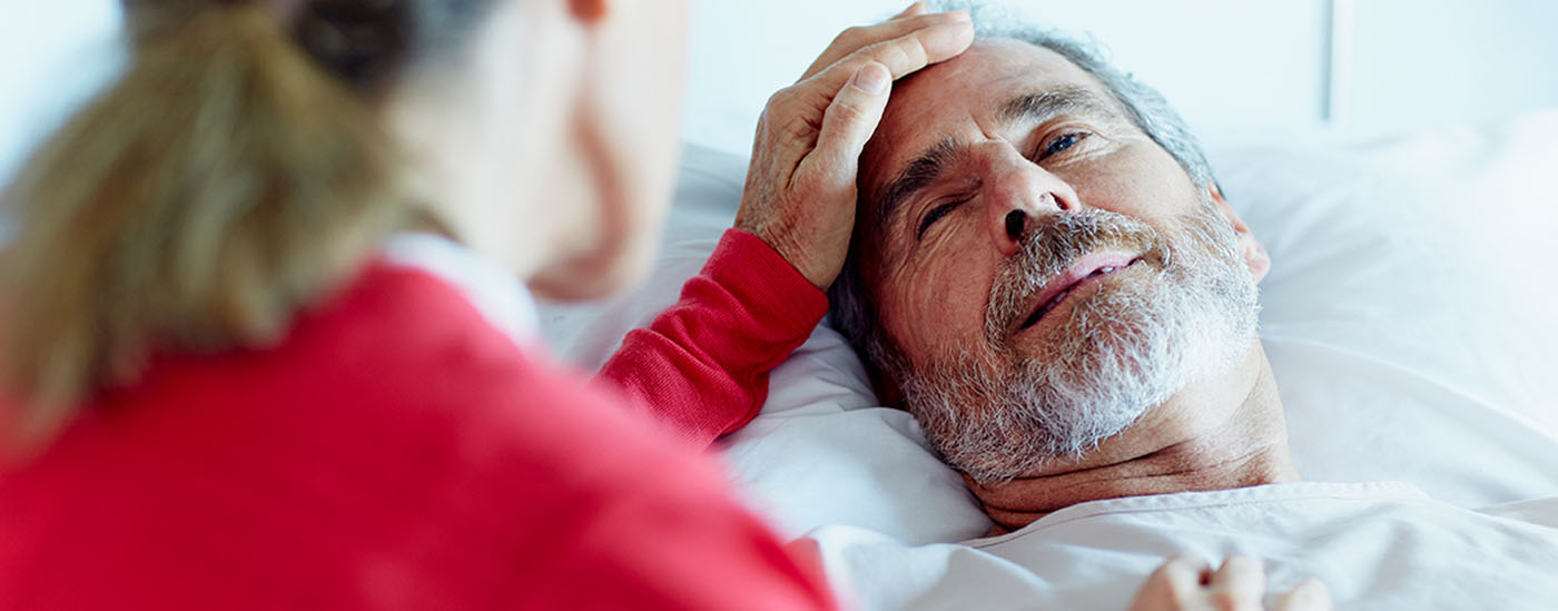 A patient lies in a hospital bed holding the hand of his caregiver