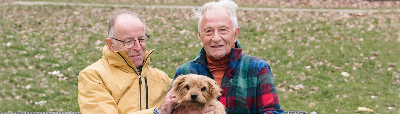 An older gay couple sits outside at the park with their dog