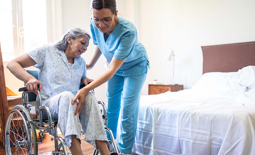 An aide assists a woman getting from bed into a wheelchair