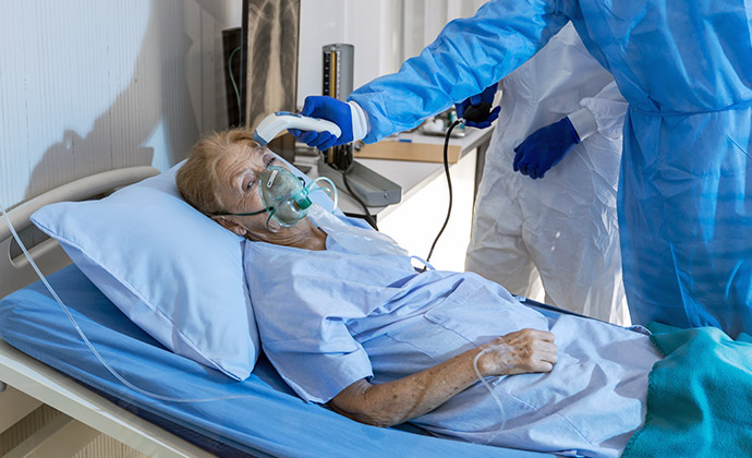 A healthcare worker takes the temperature of a woman in a hospital bed who wears a mask for supplemental oxygen