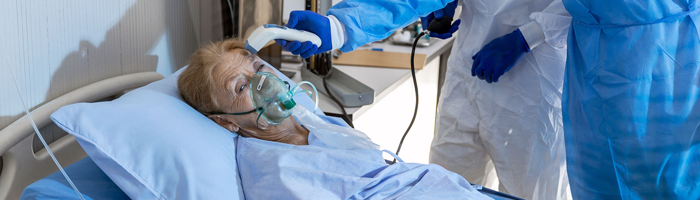 Un trabajador de servicios de asistencia médica le toma la temperatura a una mujer que yace en la cama de un hospital y que usa una máscara de oxígeno complementario
