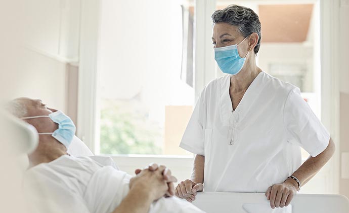 A woman in scrubs and a mask stands by a patient's bedside.