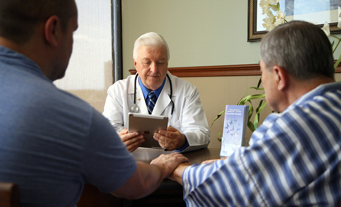 A physician consults an iPad while talking with two other men at a table