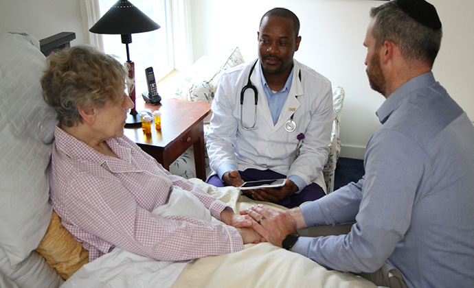 A caregiver holds the hands of a woman who is sitting up in bed, while both talk with a VITAS physician