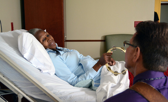 A woman lying in bed plays the tambourine while a music therapist plays guitar