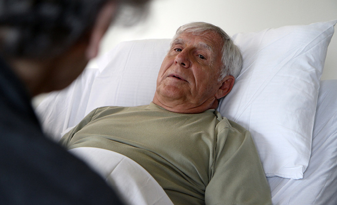 An older man talks with someone as he lies in bed