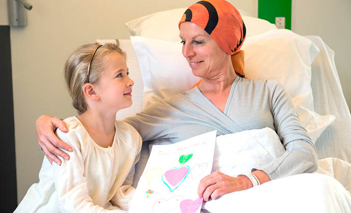 A woman sitting in bed smiles at a girl who shows her a drawing she's made