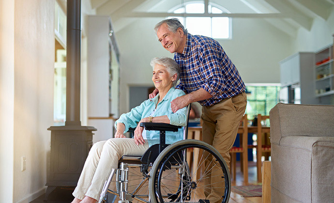 A woman in a wheelchair looks out a window, smiling, as her husband stands behind her