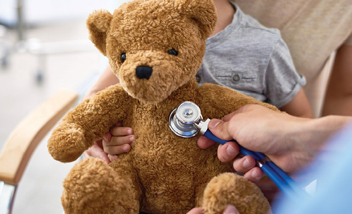 A nurse holds a stethoscope to a teddy bear, which a child is holding