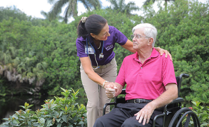 A man seated in a wheelchair talks with a VITAS team member in a tropical garden