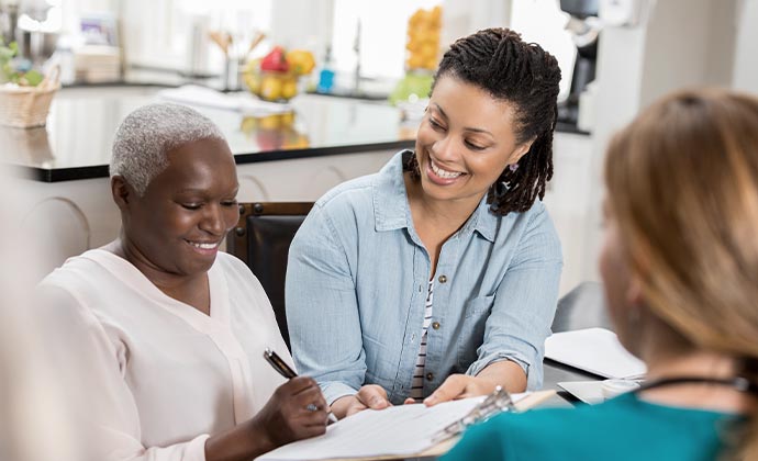 An older woman fills out a document while her daughter and a nurse look on