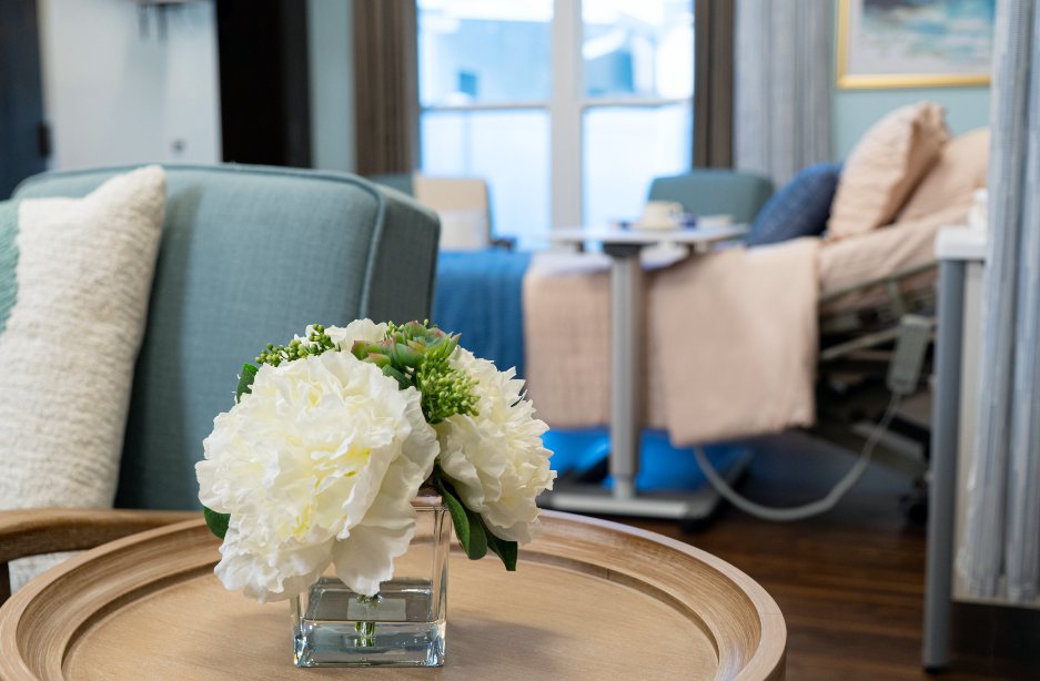 Un pequeño florero en una mesa en el frente de una habitación soleada, en el fondo hay una cama de hospital con almohadas y mantas.